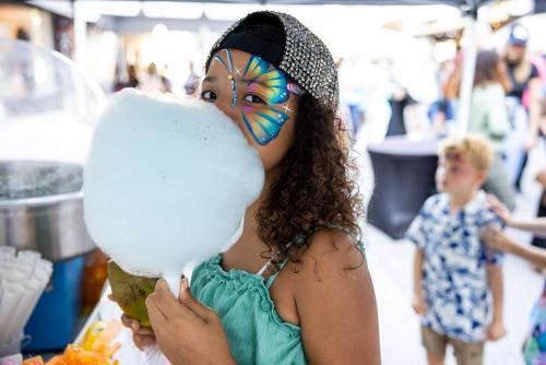 One young girl stands next to big blue cotton candy. She has butterfly painting on her cheek.