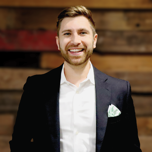 man in a white button down with black jacket over it smiles at the camera against a wood wall
