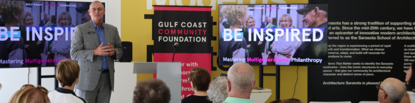 man in a gray suit stands in front of a yellow wall talking to a room full of people