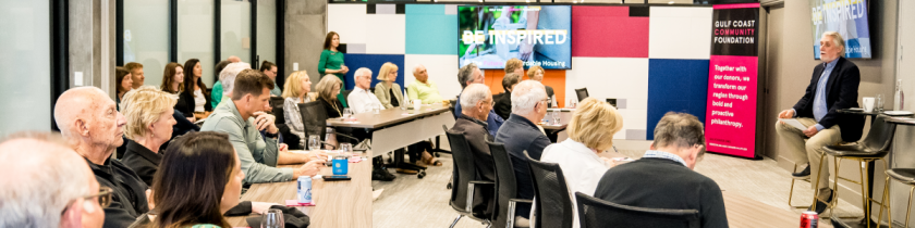 group of people sitting in a room at tables all looking at a man at the front of the room sitting in a chair talking to them