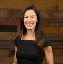 woman in a dark blue dress and dark hair smiles against a wood wall