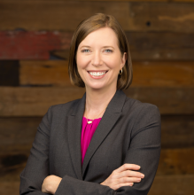 woman in a gray jacket and pink shirt smiles with her arms crossed against a wood wall