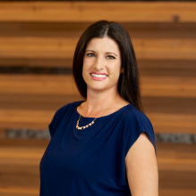 woman in a dark blue shirt and dark hair smiles against a wood wall