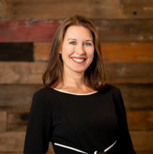woman in a black dress with white lines smiles against a wood wall