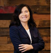 dark haired woman in a navy blue jacket with her arms crossed smiling against a wood wall