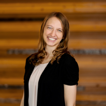 woman in a black sweater and tan shirt with light hair smiles at the camera against a wood wall