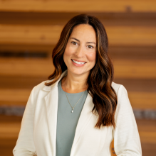 woman in a white jacket and light shirt with dark hair smiles against a wood wall