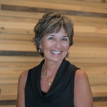 woman with short hair in a dark tank top smiles against a wood wall