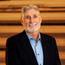 man in a blue shirt and dark jacket smiles against a wood wall
