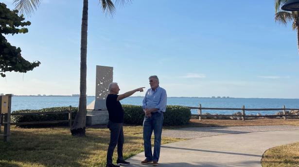 Two people standing, talking, one is pointing. Background is ocean, palm trees, sidewalk.