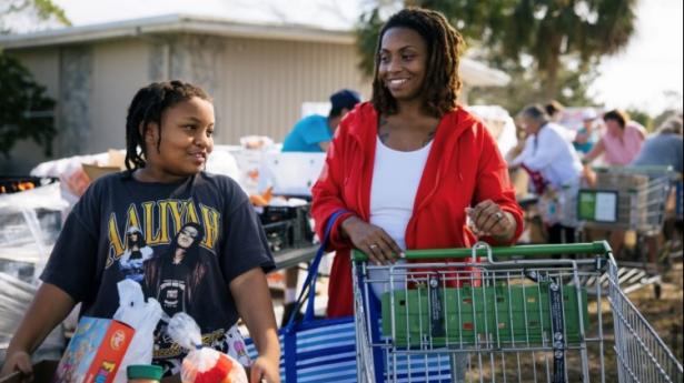 Mother and son smile together pushing grocery cart and carrying groceries.