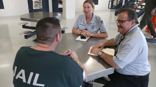 Three people sitting at table inside jail. Two people are with the Sarasota County Sheriff's Office and one person is in a jail uniform.