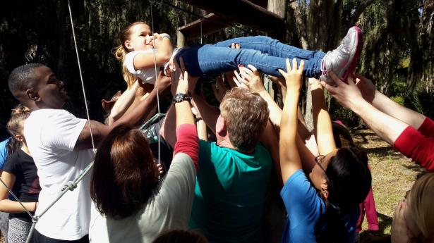 Camp Mariposa campers working together in an outdoor team-building activity focused on building resilience and trust. Students lift up girl in air, holding her up together.