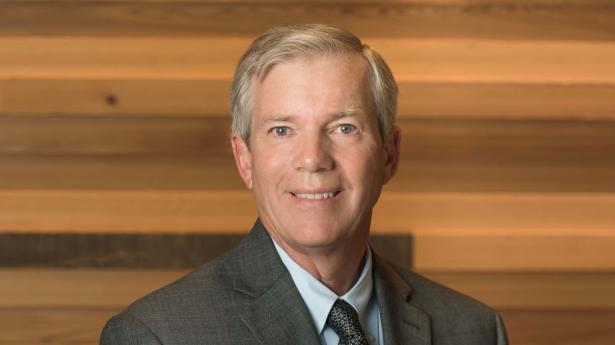 Board Chair David Sessions stands, smiling in grey suit and tie with Gulf Coast's wood wall in background.