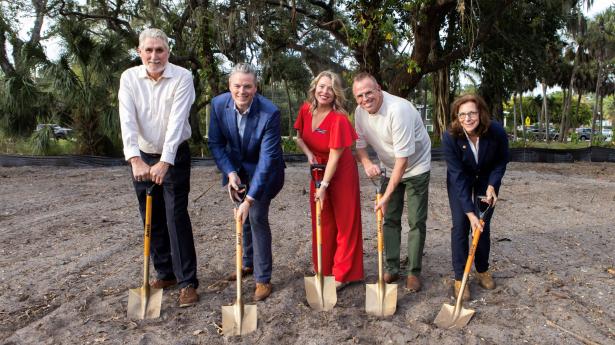 Five people stand with shovels in dirt outside smiling at camera.