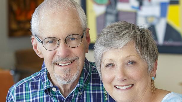 Two people, one with glasses, stand together smiling with art in background.