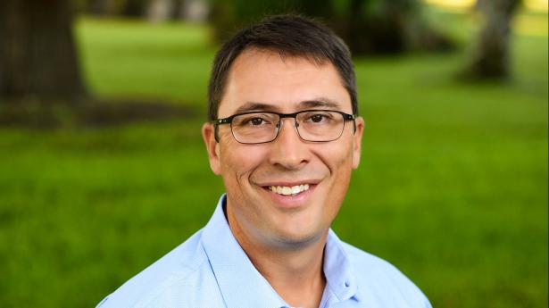 One man with brown hair and glasses stands outside smiling at camera with blue shirt on.