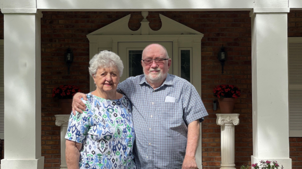 One man holding a cane and one woman stand outside of home, arms around each other.