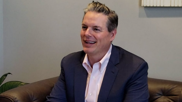 One man with dark hair in suit with pink shirt sits and smiles at camera.