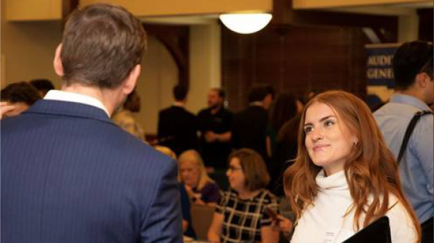 One college student with long red hair and white long sleeved shirt holds black folder and looks up at someone in conversation.