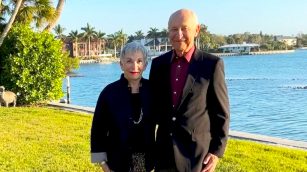 A man and woman with black jackets on smile at camera with ocean view behind them.