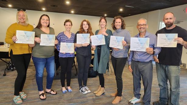 Eight adults stand indoors holding certificates and smiling.