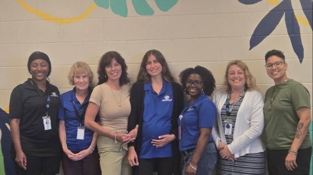 Seven people stand smiling with leaves drawn on the wall in the background.