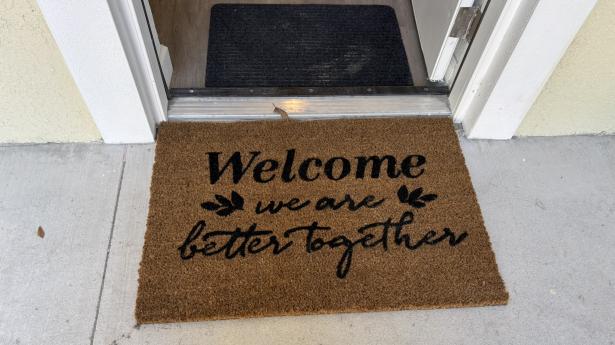 A brown mat lays on the floor of an open door saying "Welcome. We are better together."