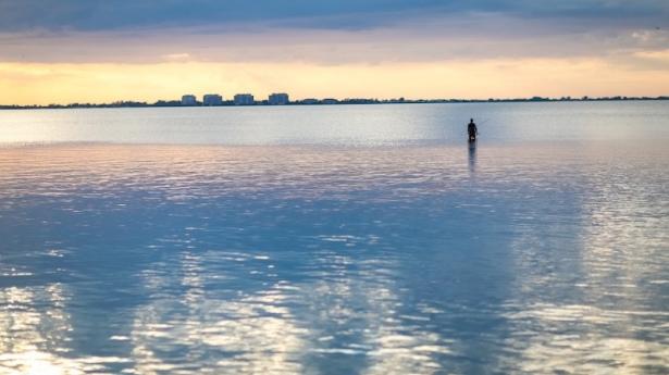 sun setting over blue waters with a cityscape in the background and the shadow of a man fishing in the water