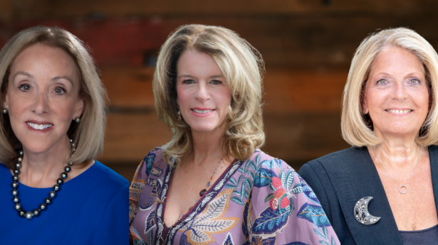 three women against a wooden wall one in blue, one in a flower dress, and one in a dark jacket