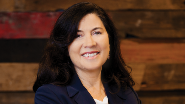 woman with dark hair in a navy blue jacket standing with her arms crossed smiling in front of a wood wall