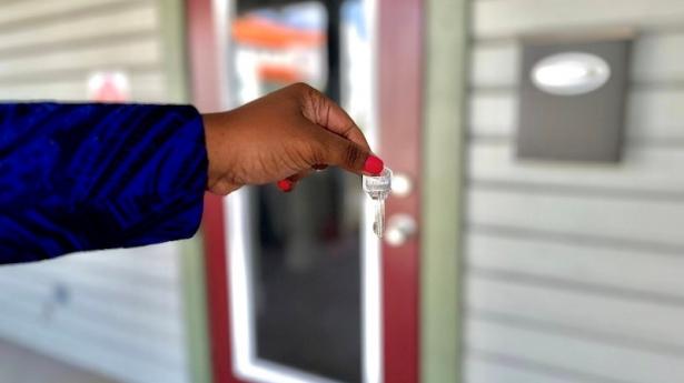 dark hand holding a silver key in front of a building with a red door