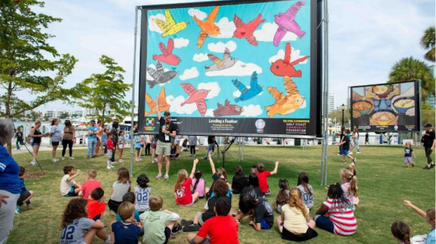 group of students sitting on the group outside looking up at a huge colorful arts piece with someone standing in front of it