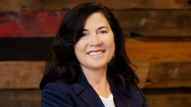 woman with dark hair stands in a dark blazer and her arms crossed against a wooden wall smiling