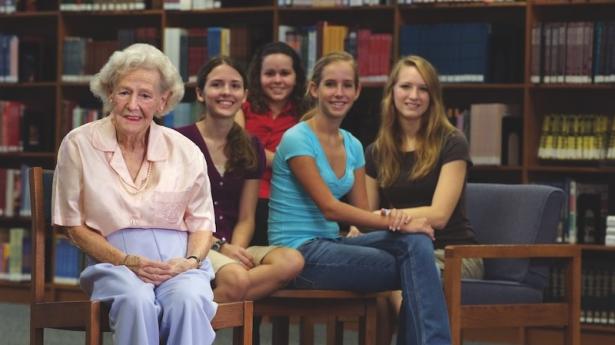 older woman sits in the library smiling with 4 young girls behind her also smiling