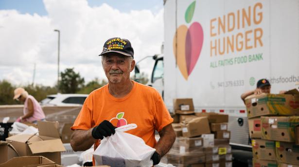 older man in a hat and an orange shirt holds a plastic bag of food outside in front of a truck that says ending hunger