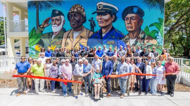 a large group of people standing outside by a mural with 4 military veterans painted on it cutting a red ribbon