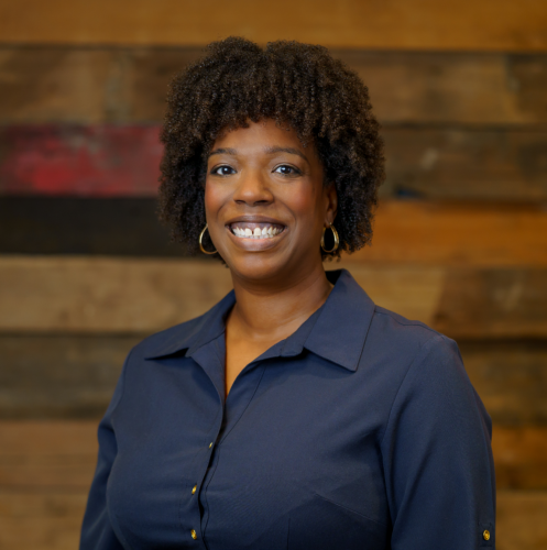 black woman in a navy blue dress smiling against a wood wall