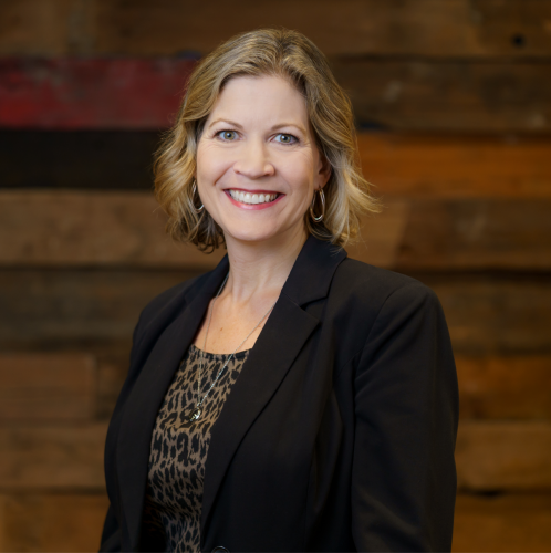 white woman with short blonde hair in a black blazer stands smiling against a wood wall