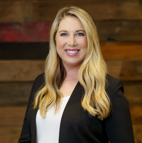 white woman with long blonde hair in a black blazer and white top stands smiling against a wood wall