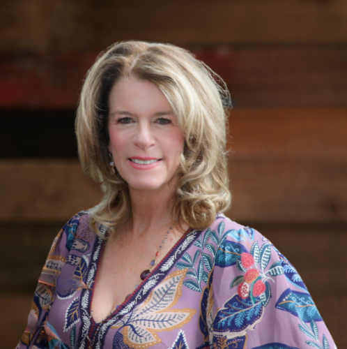 woman in a purple dress with leaves all over it and blonde hair smiles at the camera against a wood wall