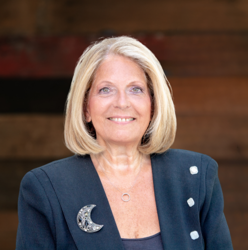 blonde woman in a navy suit smiles at the camera against a wood wall