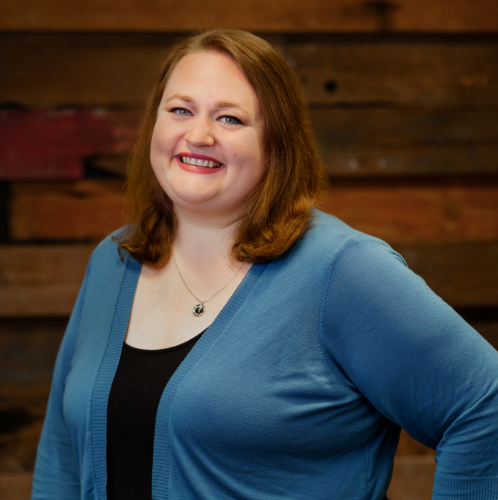 woman in a blue sweater and black dress with her hand on her hip smiles at the camera against a wood wall