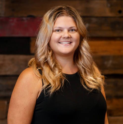 blonde woman in a black dress stands smiling against a wood wall