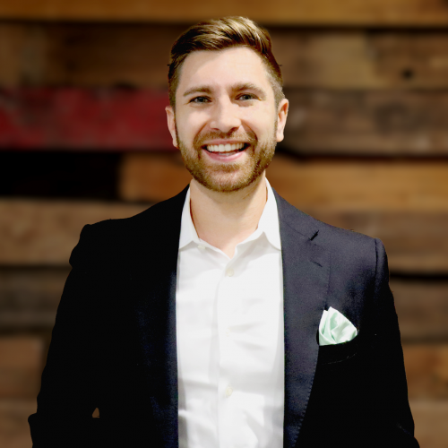 man in a white button down with black jacket over it smiles at the camera against a wood wall