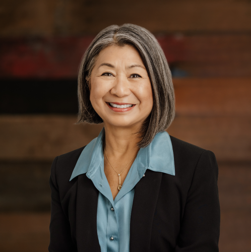 woman in a light blue shirt and black jacket smiles at the camera against a wood wall