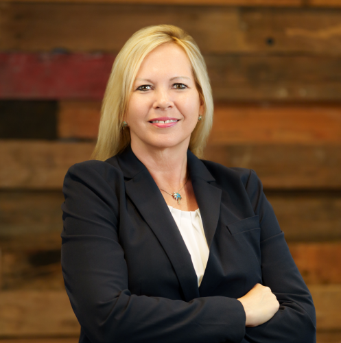 woman in a black blazer and white shirt stands with her arms crossed smiling against a wood wall