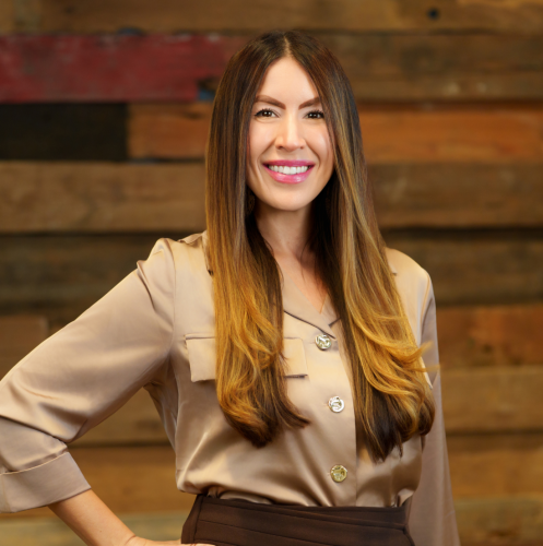 woman in a tan shirt smiles with her hand on her hip against a wood wall