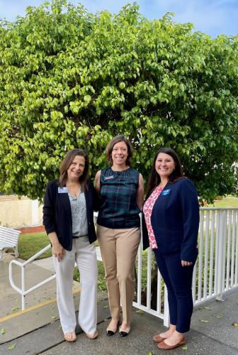 Three women stand outside by staircase and large green tree smiling.