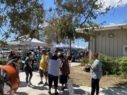 People standing outside gathering around tents with large fan in view.